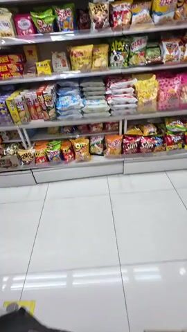 This is a video taken inside a convenience store in Daejeon, South Korea. The camera pans across several shelves stocked with a wide variety of packaged snacks and drinks. The shelves are well-organized and brightly lit, suggesting it's daytime or the store has excellent interior lighting.

The primary subjects are the inanimate objects on display: numerous bags of chips, crackers, cookies, pretzels, and other assorted snacks. The packaging features a mix of Korean and English text, with many brands and product names visible, such as "Pretzel," "Sun," and various Korean snack brands. There are also some beverages visible on the top shelf. The setting is a typical retail environment, with tiled flooring and white walls.

The activity taking place is the recording of the store's inventory, likely for promotional purposes or as a personal record. There are no people directly visible in the frame, though the perspective suggests someone is holding the camera and walking through the aisle. The overall mood is neutral and informative, focusing on the products available. The visible text is primarily product labeling and brand names, common in a retail setting.