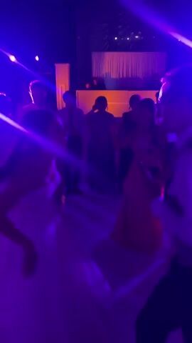 The video shows a wedding reception with the bride and groom dancing amidst other guests under purple lighting. The bride wears a white strapless gown, while the groom is in a vest and dress shirt. Several other people are visible, dressed in a mix of formal and semi-formal attire. In the background, there is a DJ booth decorated with white panels and a curtain. The dance floor is illuminated with spotlights. The scene suggests a celebratory and festive atmosphere. The camera view is shaky and at an angle, seemingly from someone also on the dance floor.
