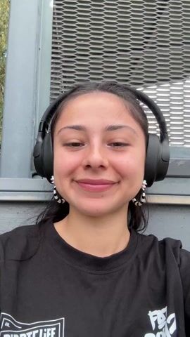 A young woman with medium-length dark hair pulled back in a ponytail smiles widely at the camera. She has large brown eyes and is wearing large black headphones over her ears. She is also wearing a black t-shirt with a white design on the front. Her earrings are light colored with dark accents. She is seated, leaning against what appears to be a grey metal structure with a grate pattern. The background is blurry, showing some greenery. The lighting suggests it is daytime outdoors.