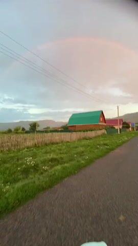 The video begins with a view of a faint rainbow arcing across a cloudy sky. A rural road stretches into the distance, bordered by green fields and scattered houses. Power lines are visible against the sky. As the video progresses, the camera pans and moves, showing different houses and more power poles. There are glimpses of a sunset with orange and pink hues in the sky. In the final moments, the camera tilts, and a young woman with her hair pulled back smiles at the camera, with a blurred background of grass and distant hills. The overall environment appears to be a rural village at dusk.