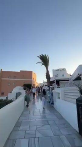 The video shows a view from a sidewalk in Oia, Santorini. People walk along a white tiled path bordered by white walls. A few buildings are visible, including one in an orange hue. A palm tree stands nearby. The camera pans to reveal a breathtaking view of the Aegean Sea and the surrounding caldera landscape. The white-washed buildings of Oia stretch along the cliffside, punctuated by the iconic blue-domed churches.