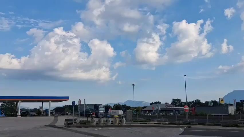 The video shows a time-lapse of a cloudy sky over a somewhat industrial or commercial area. In the foreground, there are paved areas with some planters and what appear to be barriers, suggesting a parking lot or a designated waiting area. In the background, there are buildings, possibly shops or businesses, with signage visible. A gas station canopy with blue and orange accents is present on the left. There are also streetlights and some trees scattered throughout the scene. Beyond the immediate area, distant mountains are visible under the shifting cloud cover. The sun intermittently breaks through the clouds, creating beams of light. One of the buildings in the background has visible text that appears to read "AGRO LOGISTIKA".