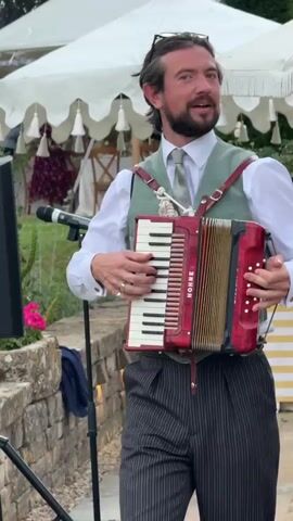 A man wearing a white shirt, grey tie, and a green vest plays a red accordion. He is standing on a stone patio in front of a white tent with decorative fringe. The man is smiling and looking to his right. In the background, a woman in a blue dress is walking away from the camera. There is a glass of orange liquid on a stone wall in the foreground. The video is about 4 seconds long.