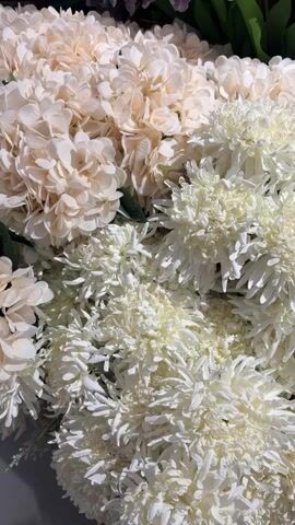 This is a close-up shot of a bouquet of artificial flowers. The bouquet contains two types of flowers. The first type are creamy white hydrangea-like flowers, which are large and clustered together. The second type are bright white, frilly flowers that appear to be chrysanthemums or similar blooms. Some green leaves are visible amongst the flowers. The lighting is bright, suggesting an indoor setting with good lighting or natural light. The video pans slowly upwards and slightly to the right, showcasing the textures and layers of the floral arrangement.