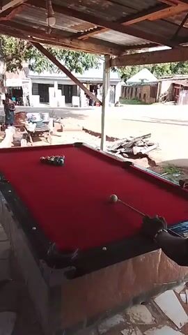 A billiard table with a red felt surface is positioned under a covered, open-air structure. Several colorful billiard balls are scattered across the table. A white cue ball is in motion towards the other balls. A hand holding a billiard cue is visible in the foreground. In the background, there are buildings, trees, and people. The scene appears to be outdoors.
