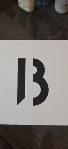 A hand opens a white box with a black "13" printed on it. The box contains a bottle of Byredo Super Cedar eau de parfum. The bottle is clear with a white label and a black cap. The box also contains a white card. The background is a dark gray surface.