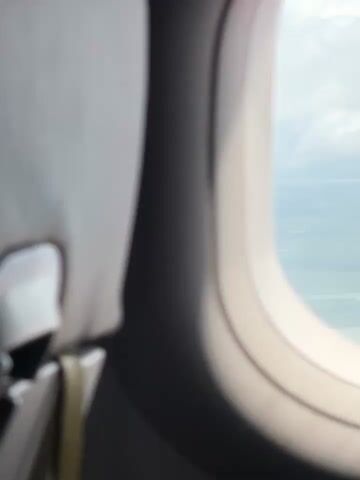 This video shows a view from an airplane window as the plane flies over islands and water. The window frame is visible on the left side of the screen, and a gray airplane seat is partially visible behind it. The view through the window shows a body of water with several islands, some of which are mountainous. Clouds are present in the sky, obscuring some of the view. The lighting suggests it is daytime, but the clouds make it appear somewhat overcast. The camera appears to be stationary, focusing on the view from the airplane.