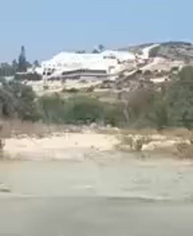 A wide shot captures a large, white, multi-story building constructed on a hillside under a clear sky. The building appears to be in a state of development or disrepair, with visible scaffolding and exposed structural elements. The surrounding landscape is dry and arid, characterized by sparse vegetation, sandy soil, and rolling hills. The foreground of the video shows a dirt road or path. The camera is stationary, and no individuals or distinct actions are observable.