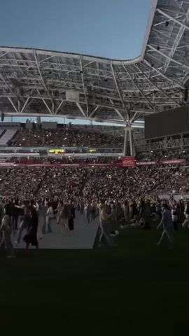 A wide shot captures a crowded outdoor stadium at dusk. The stadium features a large, modern, white-streaked roof structure and tiered seating filled with people holding up their cell phones, illuminating the scene with their lights. In the foreground, a large expanse of green grass serves as a performance or gathering area. Several people are visible on this grassy area, some standing and interacting. Two individuals wearing bright yellow, reflective safety vests with "BOSS" written on them are positioned prominently in the lower half of the frame. Behind them, metal barricades are set up, separating the front area from the main crowd. Further back, a stage with lighting and speakers is visible, with a haze or smoke effect in the air. The sky above is a clear, pale blue, indicating either early evening or late twilight. The overall atmosphere appears to be that of a large-scale event, possibly a concert or festival.