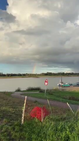 A wide shot shows a river under a cloudy sky with a rainbow visible in the distance. In the foreground, a dirt path leads toward the river, with grass and a pile of red netting on the right side. A road sign with a bicycle symbol and a warning symbol is visible in the middle right of the frame. On the river, a small boat is docked on the right side. The sun is setting, casting a warm glow on the scene.