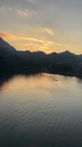 The video shows a wide river with mountains in the background, during a sunset. The sky is a mix of orange and grey. The mountains are dark. The river reflects the colors of the sky. There are two boats in the river.  The water has small ripples on the surface. There is a building near the water on the right side of the frame.  The camera appears to be stationary.