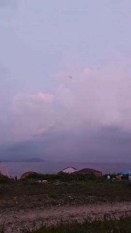 The video shows a natural landscape with a body of water in the background. In the foreground, there is a dirt path, grass, and several parked vehicles, including a silver car. There are also some tents or shelters visible near the water's edge. The sky is overcast with clouds, and the overall lighting suggests either dawn or dusk, with a soft, purplish hue. There are no prominent human activities or interactions depicted, beyond the presence of the vehicles suggesting human presence or recent activity. The environment appears to be a coastal area or a lakeshore, with land extending into the water in the distance.