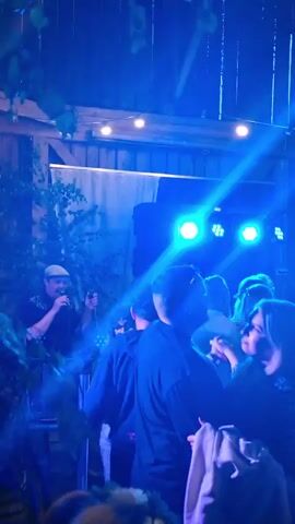 This video shows a party or event taking place inside a rustic wooden structure, possibly a barn, with exposed wooden beams and rafters visible. The lighting is dynamic, with colored spotlights, specifically blue, pink, and green, illuminating the scene and creating a party atmosphere. Strings of small lights are also hung across the ceiling. A diverse group of people are present, many of them are dancing and socializing. In the background, on a raised platform, there appears to be a DJ setup or a band with speakers and lighting equipment. Some individuals are holding drinks. The focus shifts slightly between different groups of people and the lighting effects. The overall mood appears festive and energetic.