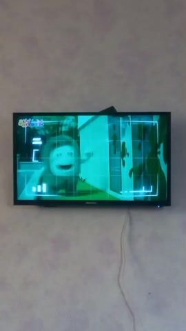 A television screen displays a scene from an animated cartoon. The cartoon features two characters, one resembling a green blob with eyes and a mouth, and the other a small robot. The background is a simplified illustration of a house with blue doors and brown splatters on the wall. The green character is on the left, gesturing with its hands. The robot is on the right, holding a yellow object. The television is mounted on a textured wall.