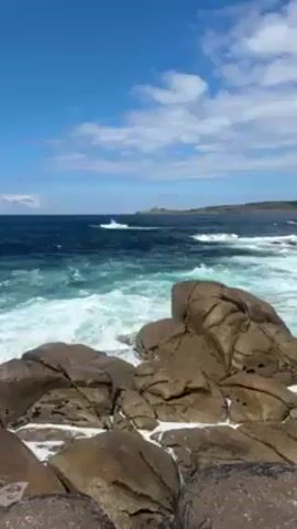 Here is a concise description of the video:

The video shows ocean waves crashing against a cluster of large, rounded rocks. The water is a mix of deep blue and turquoise, with white foam and spray from the crashing waves. The sky is bright blue with scattered white clouds. The horizon includes a headland in the distance.