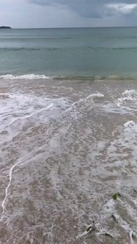 The video opens with a shot of gentle waves washing over a sandy beach. As the camera pans down, a pair of bare feet are seen standing in the wet sand near the water's edge. A pair of white sandals lies on the sand to the left of the feet, and a woven bag with "Chloe" branding is visible below them. Another pair of black slides with "NIKE" written on them are placed further to the left. The camera continues to move, revealing another set of bare feet in the sand, also appearing wet. The scene is focused on the ground and the immediate surroundings of the beach.