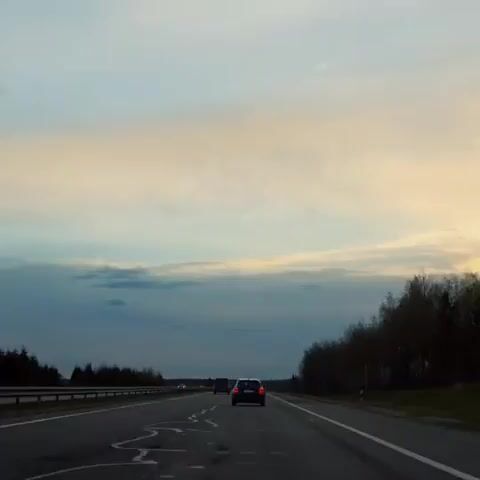 The video captures a wide shot of a multi-lane highway from the perspective of a moving vehicle. The scene depicts an open road stretching into the distance, with several cars visible ahead. The car directly in front is a dark-colored sedan. On both sides of the highway, there are dense lines of trees, appearing bare or with sparse foliage, suggesting a late autumn or early spring season. A metal guardrail runs along the left side of the road.

The sky dominates the upper half of the frame, showcasing a beautiful twilight or dawn sky. The horizon features a band of soft, light blue and pale yellow/orange clouds, indicating either sunrise or sunset. Above this band, the sky transitions to a lighter, somewhat hazy blue. Further down, darker, more solid clouds are visible. The overall lighting suggests either early morning or late evening.

The road surface shows some wear, with cracks and lighter patches visible, particularly in the foreground. White road markings, including solid lines and dashed lines for lanes, are present. The activity is simply driving on a highway. There are no people explicitly visible, nor is there any discernible text. The setting is likely a rural or semi-rural area, given the presence of extensive tree lines, specifically identified as Лошніцкі сельскі Савет, Belarus.