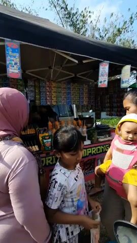 A group of people, including two adult women and two children, are gathered in front of a food stall. One woman, wearing a pink hijab, has a baby strapped to her chest in a pink carrier. The baby is wearing a yellow hood. Another woman, also wearing a hijab, is standing to the left. A young girl with dark hair is looking down at something in her hands. The food stall has a black awning and is stocked with drinks and snacks. There are several bottles of what appear to be drinks and some fried food items on display. The environment appears to be outdoors, possibly a market or street vendor setting, with trees visible in the background. The lighting suggests it is daytime.