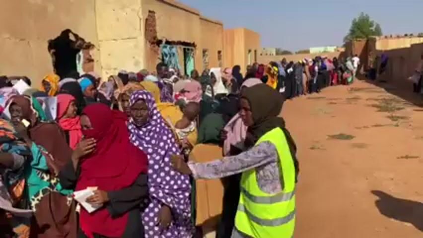 A crowd of people stand in a long line on a dirt road between buildings. Most of the people appear to be women wearing hijabs of various colors and patterns. A few children are present. A person wearing a neon yellow vest appears to be directing the line. A man in a plaid shirt and backpack walks past the line, looking at the camera. The buildings are simple, single-story structures, and some appear to be in disrepair. The setting looks like a rural or impoverished area. The weather is sunny and the sky is clear. The general atmosphere seems to indicate people are waiting for something, possibly aid or a service.