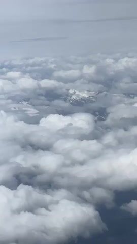 The video shows an aerial view of a landscape covered in clouds. The camera angle is looking down from above, with thick white clouds filling most of the frame. In the distance, peeking through gaps in the clouds, are snow-capped mountains. The sky above the clouds is a pale gray. The overall impression is one of a vast, serene, and somewhat majestic natural landscape.