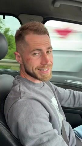 A man with short brown hair and a beard is driving a car. He is wearing a gray sweatshirt and a seatbelt. A person in a light blue denim jacket is in the passenger seat, patting the man's head. The man is smiling and laughing. They are driving on a highway with greenery visible through the windows.