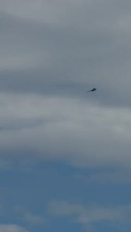 A dark silhouette of a jet fighter is captured against a cloudy blue sky. The jet appears to be in mid-flight, with its wings extended and its nose pointed slightly downward. The clouds are a mix of white and gray, with some areas appearing darker than others. The overall lighting suggests it is daytime, possibly under an overcast sky. The jet is a small, distant object in the frame, suggesting it is far away. The video is a still image that appears to be a single frame from a video, as indicated by the time stamp at the beginning of the input.