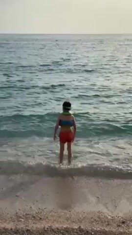 A video captures a child wading in the ocean at the shoreline. The child is wearing red shorts and a blue-green flotation device.  A man, also in red shorts, throws a pink pool noodle towards the child. The camera then pans up to show the sun setting over the water, creating a shimmering reflection on the surface. The sky appears partially cloudy.