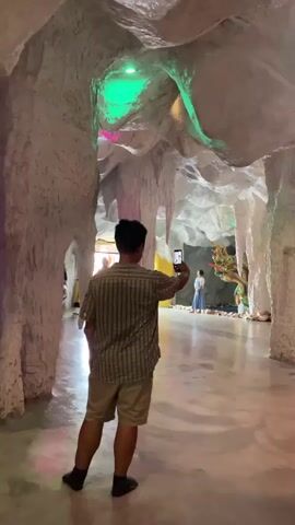 A man is filming a statue inside a cave-like structure. The statue appears to be a seated figure in a white outfit, adorned with beads and jewels, positioned on a throne-like structure. To the right of the statue, a colorful serpent-like carving is visible. The cave walls are textured and rough, with stalactite-like formations hanging from the ceiling. Green and pink lights illuminate the cave, casting colorful reflections on the polished floor. The man in the foreground is wearing a plaid shirt and shorts and is holding his phone up to record. Another person can be seen in the background, further down the cave passage.