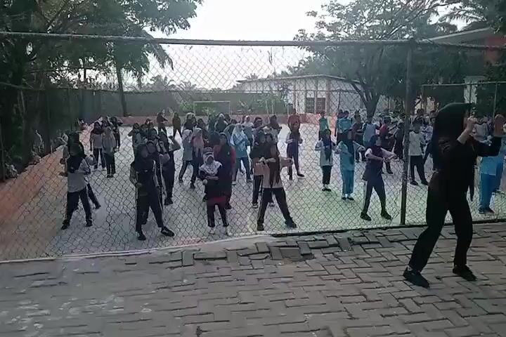 A group of people, mostly children and young adults, are exercising outdoors. They are wearing casual clothes and some of the girls are wearing hijabs. They are in a fenced-in area, possibly a schoolyard or sports complex. Some adults are also present and appear to be supervising or participating in the exercise. The ground is paved with bricks. The overall atmosphere appears to be active and energetic.
