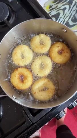 The video shows donuts frying in a pot of oil on a stovetop. There are six donuts in the pot, and they are golden brown and puffy. The oil is bubbling around the donuts, indicating that they are cooking. The pot is silver and appears to be made of stainless steel. The stovetop is black and appears to be gas. There is a red rug visible in the lower right corner of the frame.