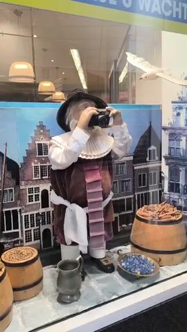 A mannequin of an old man with a white beard and a black hat stands in a shop window. He is wearing historical clothing, including a ruffled white collar, a maroon vest with pink ties, and a white sash. The mannequin holds a black camera to his face, appearing to take a picture. The background of the window display depicts a European city street scene with old brick buildings. To the right of the mannequin, there are two wooden barrels, one filled with brown cylindrical objects, and a smaller bowl containing blue objects. To the left, another wooden barrel is visible. A metallic jug sits on the ground in front of the mannequin. A white model bird, resembling a seagull, is positioned in the upper right corner of the window. The window is lit from above by modern ceiling lights, and the reflection of these lights is visible on the glass. A blue and yellow striped banner is partially visible at the top of the window.
