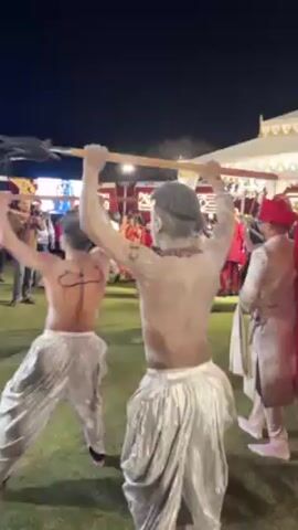 The video shows a group of men dancing, shirtless, in a field at night. The men are covered in what appears to be white paint or powder and wear silver-colored pants. They are holding wooden sticks and moving them rhythmically. In the background, there is a crowd of people, tents, and decorative lighting. The scene appears to be part of a celebration or event.