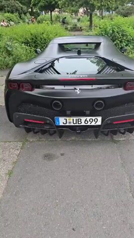 This is a rear-view, panning video of a matte black Ferrari SF90 Stradale parked on a paved surface. The car is the primary subject. Its distinctive rear design, including the quad exhaust pipes, sharp diffuser, and iconic Ferrari badge, is prominently displayed. The license plate reads "J UB 699" and has German plates with the EU star symbol. Red accents are visible on the rear brake calipers and within the diffuser grilles.

The setting appears to be an outdoor area with lush green foliage and trees in the background. There are several people visible in the background, some sitting on benches, and a man standing near a trash bin. The overall atmosphere suggests a casual daytime scene, possibly in a park or a public space in Jena, Germany, given the license plate format and the visible graffiti on a wall in the background. The lighting indicates it is daytime, likely overcast or late afternoon, as there are no strong shadows. The video captures the car from a low angle, emphasizing its aggressive stance.