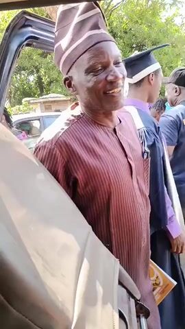 The video shows a man wearing a traditional patterned cap and a striped shirt, smiling and holding a pamphlet. He is standing next to a car, with other people in the background, some of whom are wearing graduation gowns and caps. The man raises his hand and waves the pamphlet. The scene appears to be outdoors, with trees and buildings visible in the background. The lighting suggests it is daytime.