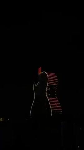 The video shows the Hard Rock Hotel in Hollywood, Florida at night. The building is shaped like a giant guitar and is illuminated with colored lights. The lights change from red to white to blue. The sky is black, and the surrounding area is dark.