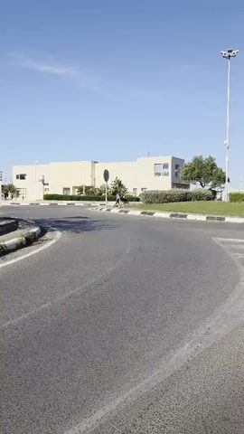A camera pans across a paved road leading to a beach. The road has painted markings and a roundabout with greenery. In the background, a light-colored building stands, partially obscured by trees. As the camera pans, the beach comes into view, with people visible along the shoreline. The sky is clear and blue. The road curves to the right, lined with vegetation on one side.