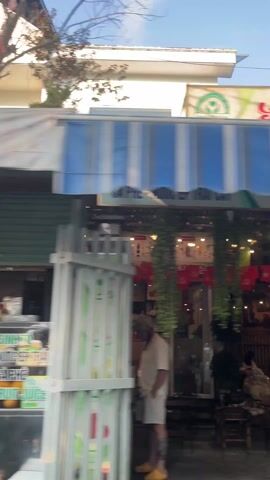 The video shows a quick pan of roadside businesses and buildings from a vehicle, likely a car or motorcycle, moving along a street. The first business observed is a food or beverage stand, "TAM," displaying fresh fruits like avocados. The next is a two-story building, "CỬA HÀNG ĐẠT TẤN," with an open-air retail area underneath, selling plants and gardening supplies. The sky is visible in the background.