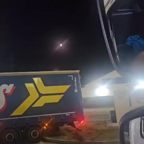 The video, recorded at night, captures a side view of a blue and yellow truck trailer. The moon is visible in the dark sky, slightly off-center and emitting a bright glow. The truck has a logo with the letters "S" in red and a yellow symbol. Lights from nearby structures are also visible. The camera appears to be inside another vehicle, as evidenced by a portion of a car interior in the foreground. The lighting is dim, creating a shadowy atmosphere.