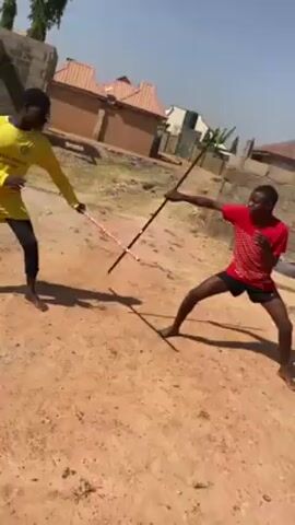 Two young men are engaged in a mock sword fight on a dirt ground outdoors. One man is wearing a yellow long-sleeved shirt and black leggings with red accents, while the other is in a red patterned t-shirt and black shorts. Each is holding a long, thin stick, using it as a pretend sword. They are both barefoot and actively moving, attempting to strike each other with their sticks. The background shows a neighborhood scene with houses and a stone wall under a bright, sunny sky. The camera angle is slightly tilted and appears to be handheld, capturing the action from a dynamic perspective.
