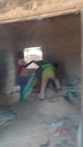 Two individuals are in an indoor environment with rough, unfinished walls and a sandy floor. One person, wearing a red and white striped shirt and blue pants, is holding a large, light-colored bag. The other person, wearing a green shirt and light-colored pants, is facing away from the camera and appears to be interacting with a piece of machinery or equipment. Bags of material, likely sand or cement, are scattered around the floor and to the left of the frame. A small window with bars is visible in the background. The lighting suggests it is daytime.