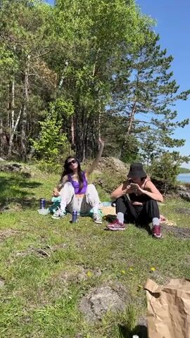 Two young women are relaxing outdoors in a natural, sun-drenched setting. The scene appears to be a grassy area with scattered rocks, bordered by a forest of pine and birch trees. A body of water, likely a lake or large river, is visible in the background under a clear blue sky.

The woman on the left is wearing a purple crop top, light-colored pants, and white sneakers, with sunglasses on her head. She initially poses with one arm raised and then settles into a cross-legged sitting position on a light blue blanket, looking forward. Two blue cans are placed near her.

The woman on the right is wearing a black bucket hat, a black crop top, and black pants, with dark-colored sneakers. She is seated on a yellow blanket and is engrossed in her smartphone.

In the foreground, to the lower right, a brown paper bag is visible. The ground is covered in green grass with small yellow wildflowers. The overall atmosphere suggests a pleasant, sunny day, with both individuals enjoying leisure time in nature. No discernible text is visible on any objects or clothing. The location context indicates Sliznevo, Russia.
