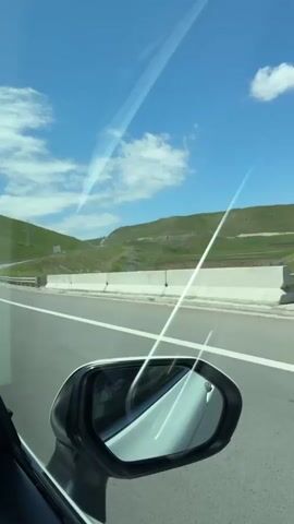 The video captures a scenic drive through the Armenian countryside, likely near Dzorakap. The viewpoint is from inside a vehicle, looking out the side window and through the side mirror. The weather is clear and sunny, with a bright blue sky dotted with fluffy white clouds. The landscape is characterized by rolling green hills, indicating a temperate season, possibly spring or summer.

The main subjects are the road and the surrounding environment. The road appears to be a well-maintained highway with clear white lane markings. A guardrail runs along the side of the road, separating it from the grassy slopes. In the distance, power lines and poles can be seen, suggesting infrastructure development in the area.

The primary activity is driving. The perspective shifts slightly as the vehicle moves, offering different angles of the scenery. A white car is visible ahead, traveling in the same direction on the highway. The reflection in the side mirror shows the interior of the car, including a portion of the dashboard and the side window frame.

Notable details include the sunlight creating streaks of light across the video, likely due to reflections on the car's windows or lenses. The time of day appears to be daytime, given the bright sunlight and clear skies. The overall mood conveyed is one of peaceful travel and appreciation for the natural beauty of the Armenian landscape. There is no visible text within the frames of the video itself, aside from what might be on the license plate of the car ahead.