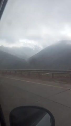 The video shows a view from a car driving on a road, with misty mountains in the background. The road has a metal guardrail along the side and a yellow line. The sky is overcast and the lighting is dim. A pole stands beside the guardrail. The side mirror of the car is visible in the lower left corner of the frame. The ground between the road and guardrail is a mix of dirt and gravel. The mountains in the background are shrouded in fog, partially obscuring their details.