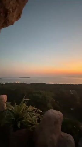 This is a vertical video captured from a high vantage point. The scene shows a landscape at dusk or dawn. The sky transitions from a pale blue at the top to a gradient of orange and yellow near the horizon, indicating either sunrise or sunset over a body of water. Below the sky, a dark, tree-covered landmass stretches across the midground, with some structures or lights visible in the distance. In the foreground, parts of a rocky outcrop and some foliage are visible, framing the view. The lighting is dim, suggesting low-light conditions typical of twilight. There are no people or explicit actions depicted in the video.