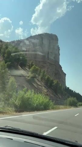 The video shows a view from inside a vehicle looking out at a large rock formation on the left side of a highway. The rock formation is steep and layered, with some vegetation growing on its slopes. The highway is paved and has white lane markings. The sky is blue with some white clouds. The sun appears to be shining. The vehicle is moving forward.