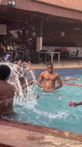 The video shows a group of shirtless young men enjoying themselves in an outdoor swimming pool. They are splashing, throwing a ball, and generally having fun. The pool is surrounded by a tiled deck, and there is a covered area with tables and chairs visible in the background. People are seated at the tables. The pool is part of an outdoor entertainment area attached to a building.