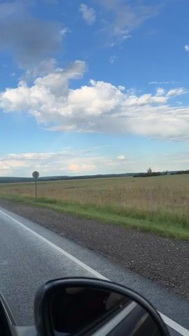 The video shows a view from inside a vehicle, looking out towards a rural landscape. The road is wet, indicating recent rain. In the distance, there is a vast field of dry grass under a bright blue sky with scattered white clouds. A faint rainbow is visible in the sky. A single circular road sign stands on the side of the road. The camera angle suggests the vehicle is moving.