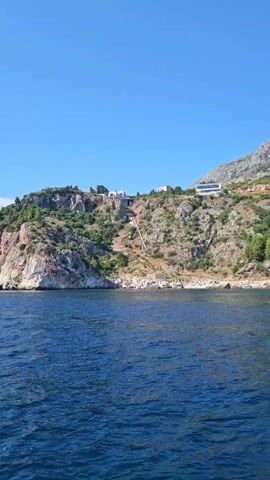 A drone shot captures a serene blue ocean meeting a rocky, green-covered cliffside. Modern white buildings are perched atop the cliff, overlooking the water. A visible pathway, likely stairs or a ramp, ascends the steep incline towards the structures, suggesting access or construction. The sky is a clear, vibrant blue, indicating a sunny day. The scene evokes a sense of luxury and isolated living.