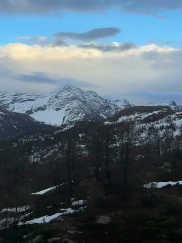 The video presents a panoramic view of a mountainous landscape, likely at dusk or dawn, with the camera slowly panning from right to left. Snow-capped peaks dominate the background under a sky with both blue patches and dramatic, light-kissed clouds. The foreground features a steep, forested slope with numerous bare trees, interspersed with patches of snow on the ground. As the camera pans, a bright, full or near-full moon becomes visible high in the sky, positioned above the distant mountains. The overall scene suggests a cold, high-altitude environment, consistent with an alpine region like Poschiavo, Switzerland.