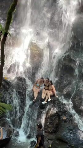 Three women in bikinis are sitting on wet, dark rocks in the middle of a cascading waterfall. The waterfall is surrounded by lush green foliage and trees, and bright sunlight is visible through the falling water. The women are posing, initially sitting still, then raising their arms excitedly. Below them, a man wearing a dark t-shirt and a cap stands on a rocky path, looking up at them, seemingly taking a picture or video. The scene appears to be set in a tropical environment, fitting with the provided context of Gianyar, Indonesia. The atmosphere is joyful and adventurous, with the women enjoying the natural beauty of the waterfall.